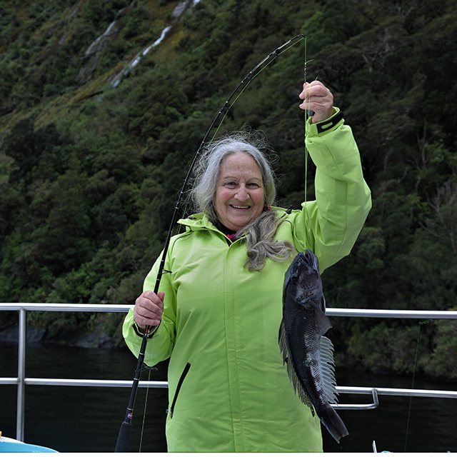 Woman in a green jacket proudly holding a fishing rod with a Blue Cod attached that she has caught.